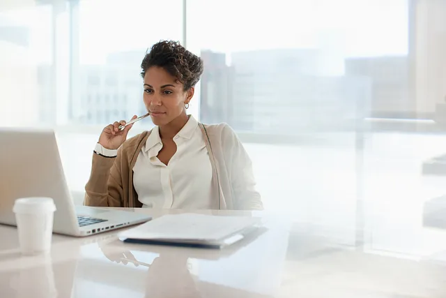 Professional woman working thoughtfully at her laptop in a bright, modern office with a coffee cup and notebook on the desk.